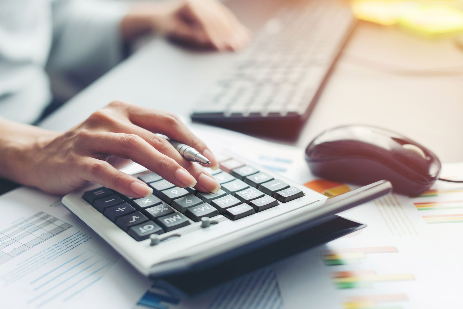 A person is typing on a calculator next to a computer mouse.