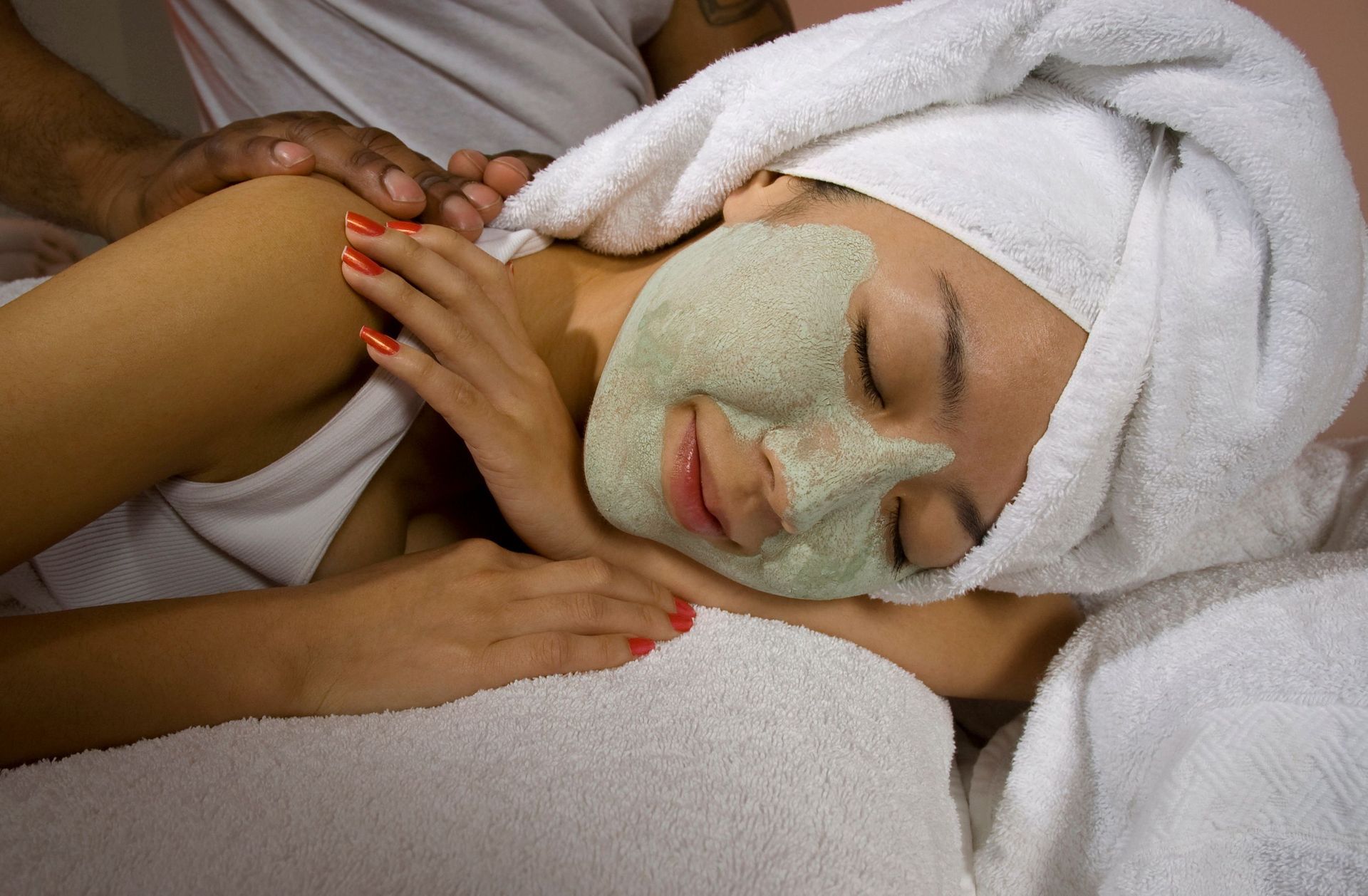 Person receiving a facial mask and head wrap during a spa treatment, resting on a towel-covered bed