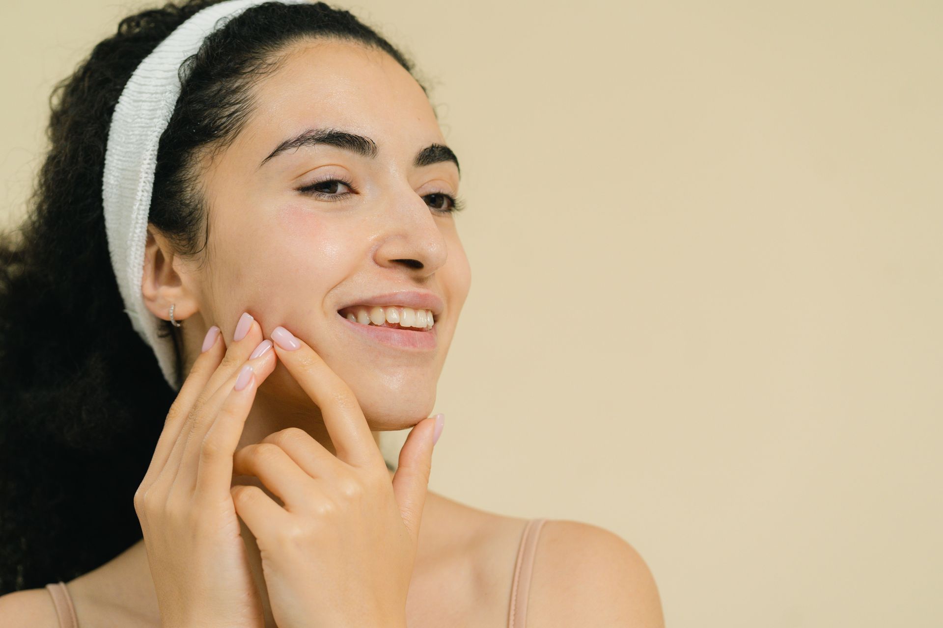 Woman applying skincare cream to her cheek, smiling against a beige background