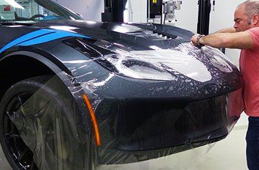 Un homme applique un film transparent sur l'avant d'une voiture de sport noire pour protéger sa peinture. La voiture est dans un garage.