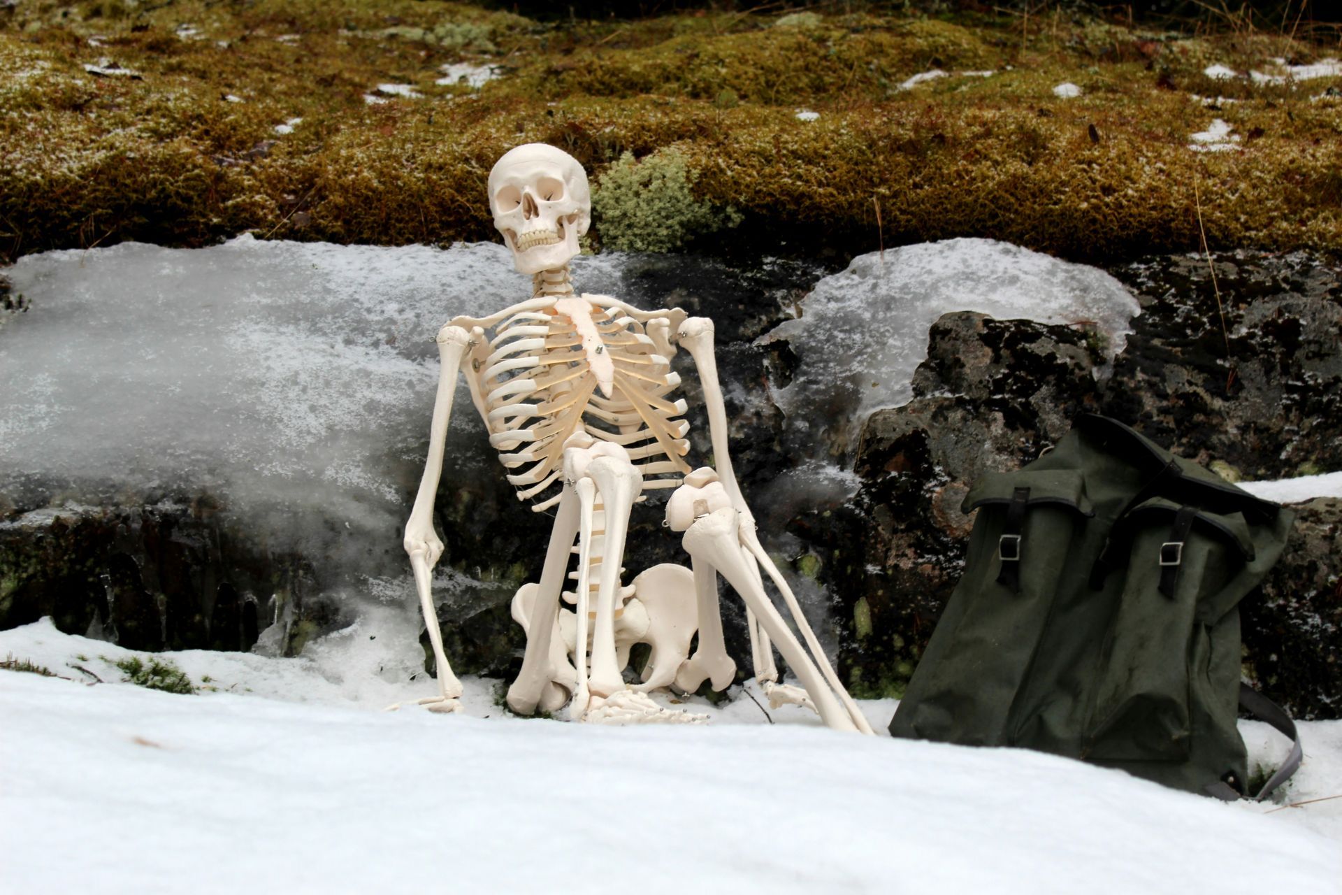 A skeleton is sitting in the snow next to a backpack