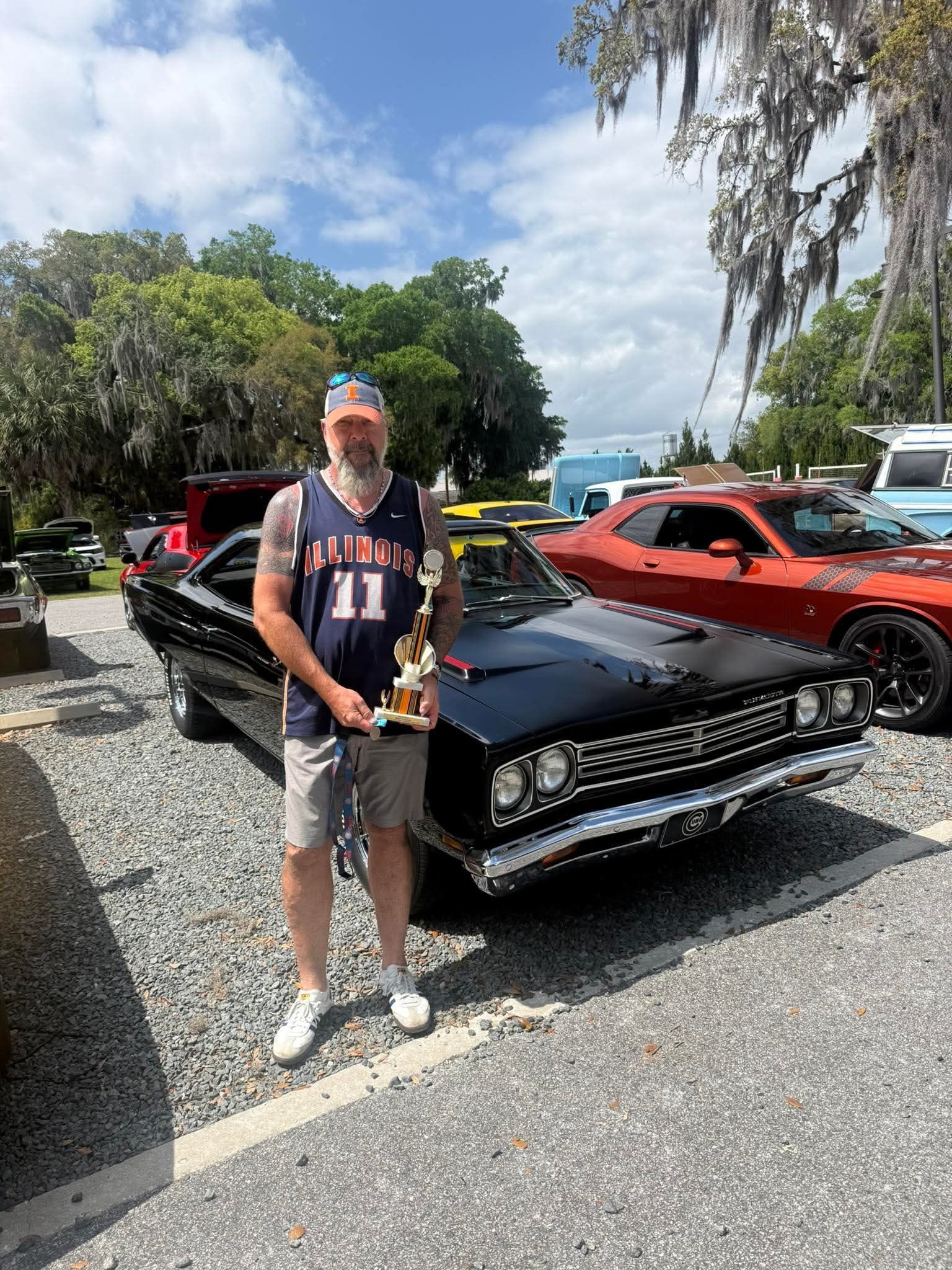 Owner standing beside a black restomod muscle car holding a trophy at a Florida car show