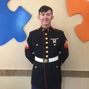 Marine in dress blues uniform, smiling, standing in front of puzzle-shaped wall decor.