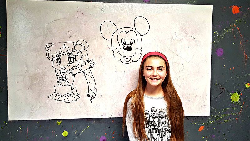 Girl stands near drawings of Sailor Moon and Mickey Mouse on a white board.