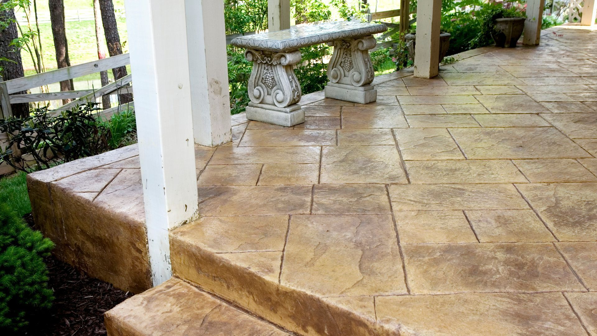 A stamped concrete patio with decorative bench, steps, and white columns, in a yard setting.
