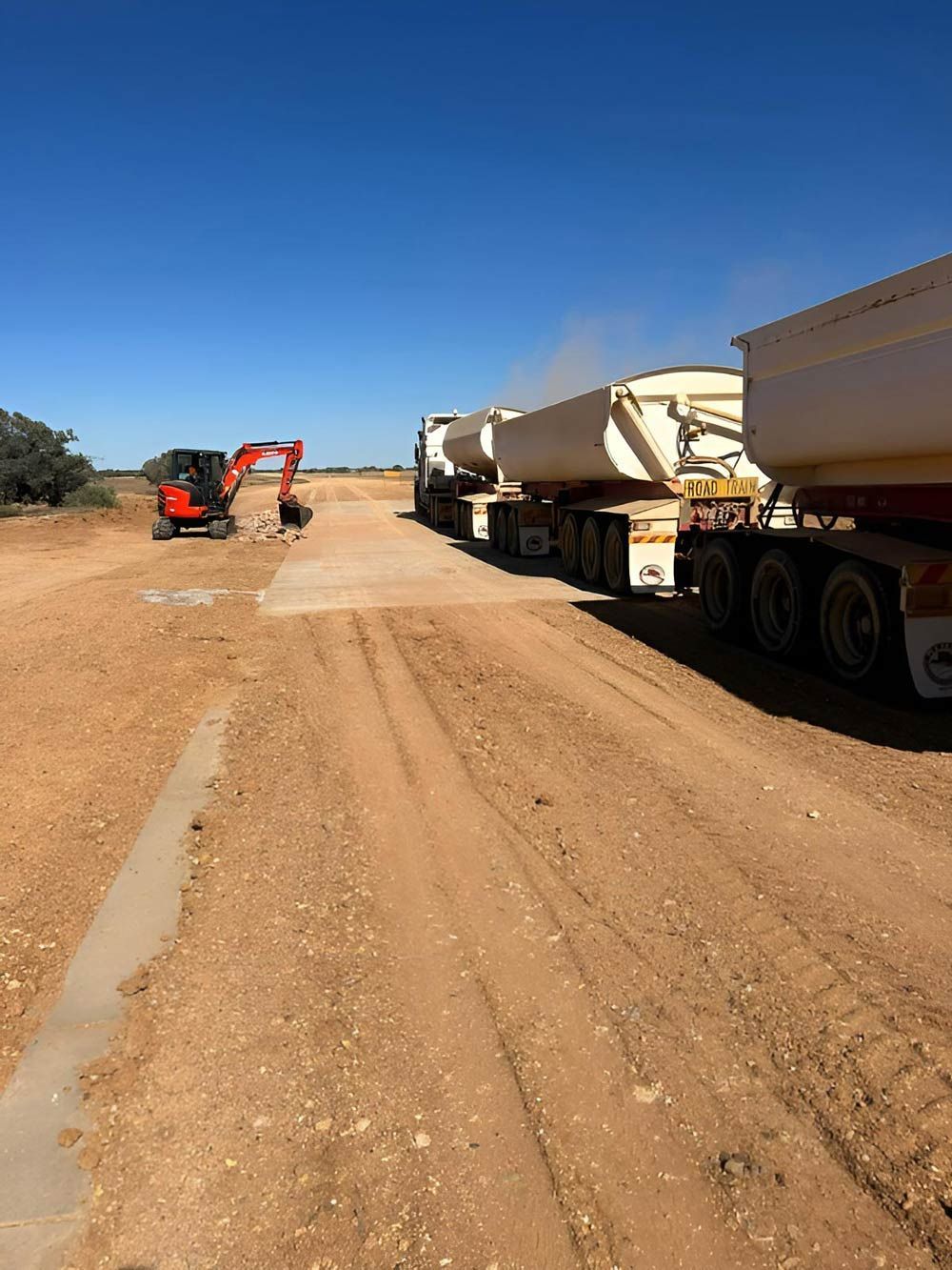 A Row Of Semi Trucks Are Parked On A Dirt Road With Excavator — NQES Industries in Julia Creek, QLD