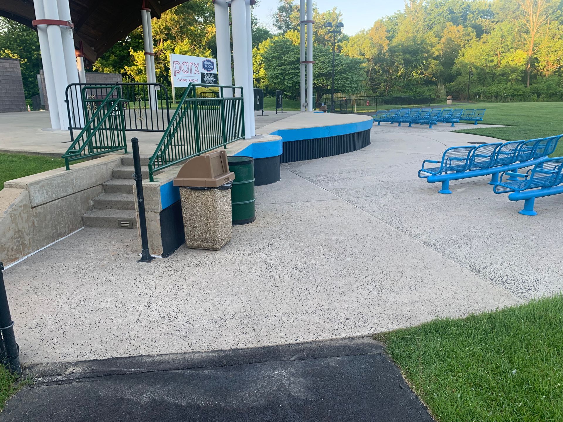 Park stage with concrete surface, blue seating, trash cans, and green railings. Trees in the background.