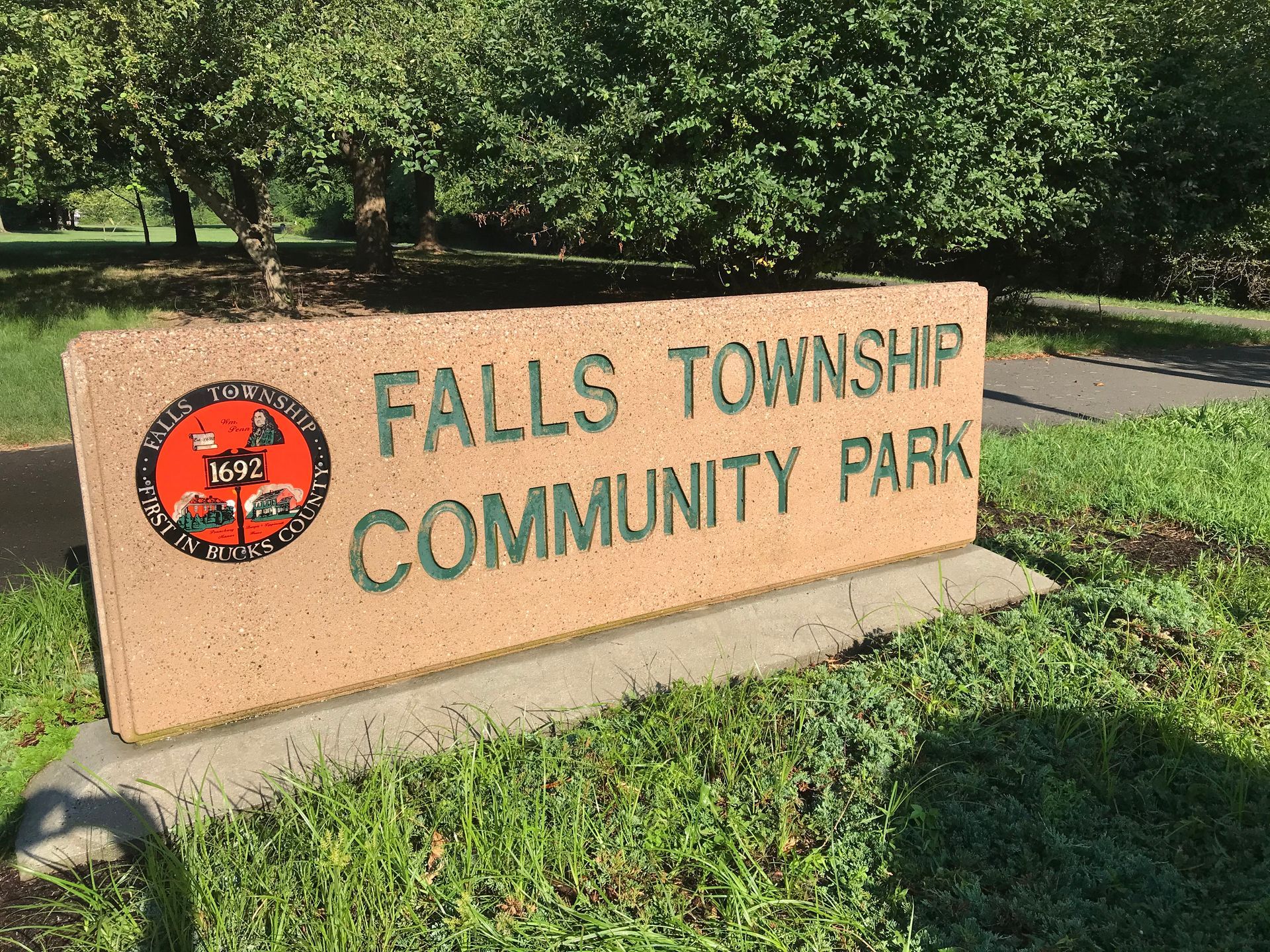Sign for Falls Township Community Park; beige stone with green lettering, logo on left, trees and path in background.