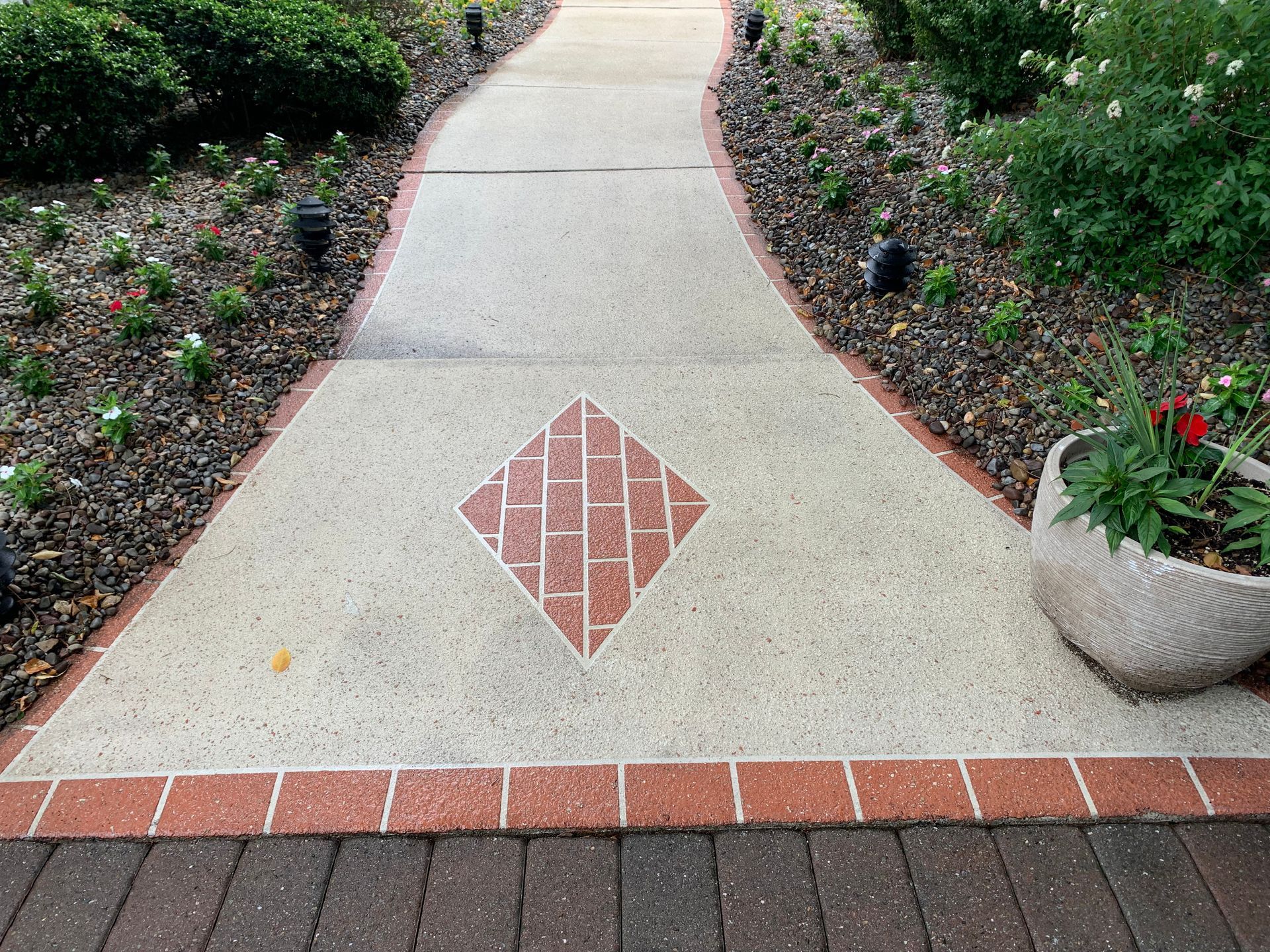 Concrete walkway with red brick border, diamond-shaped brick inlay, surrounded by landscaping.