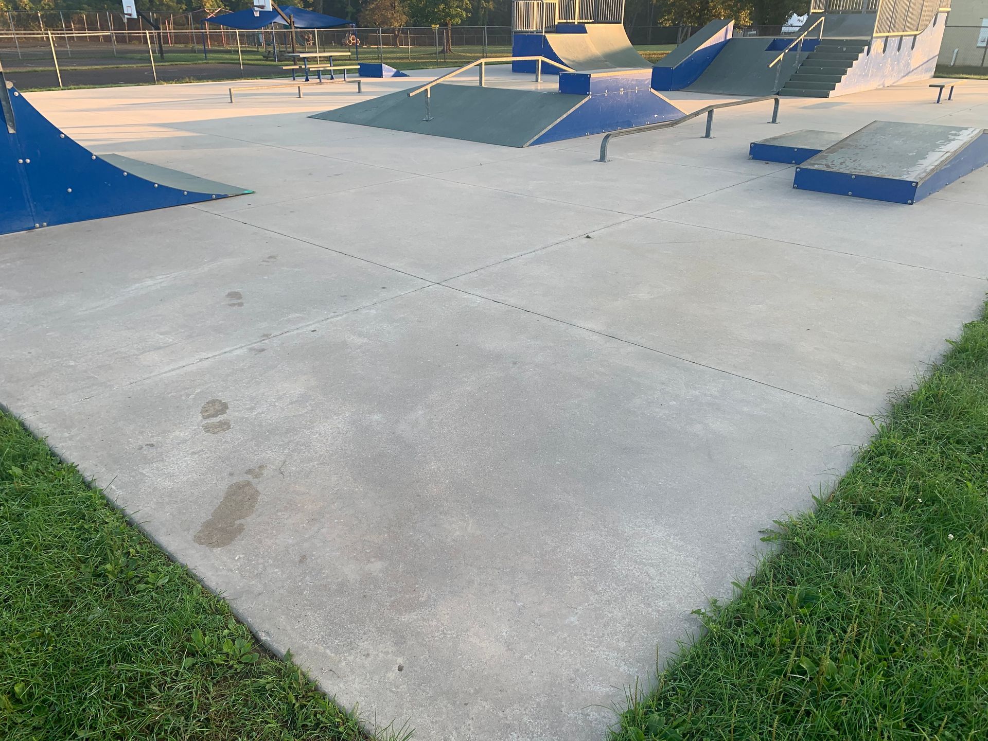 Skatepark with concrete ramps and rails. Blue, green, and gray structures sit on the concrete. Green grass surrounds it.