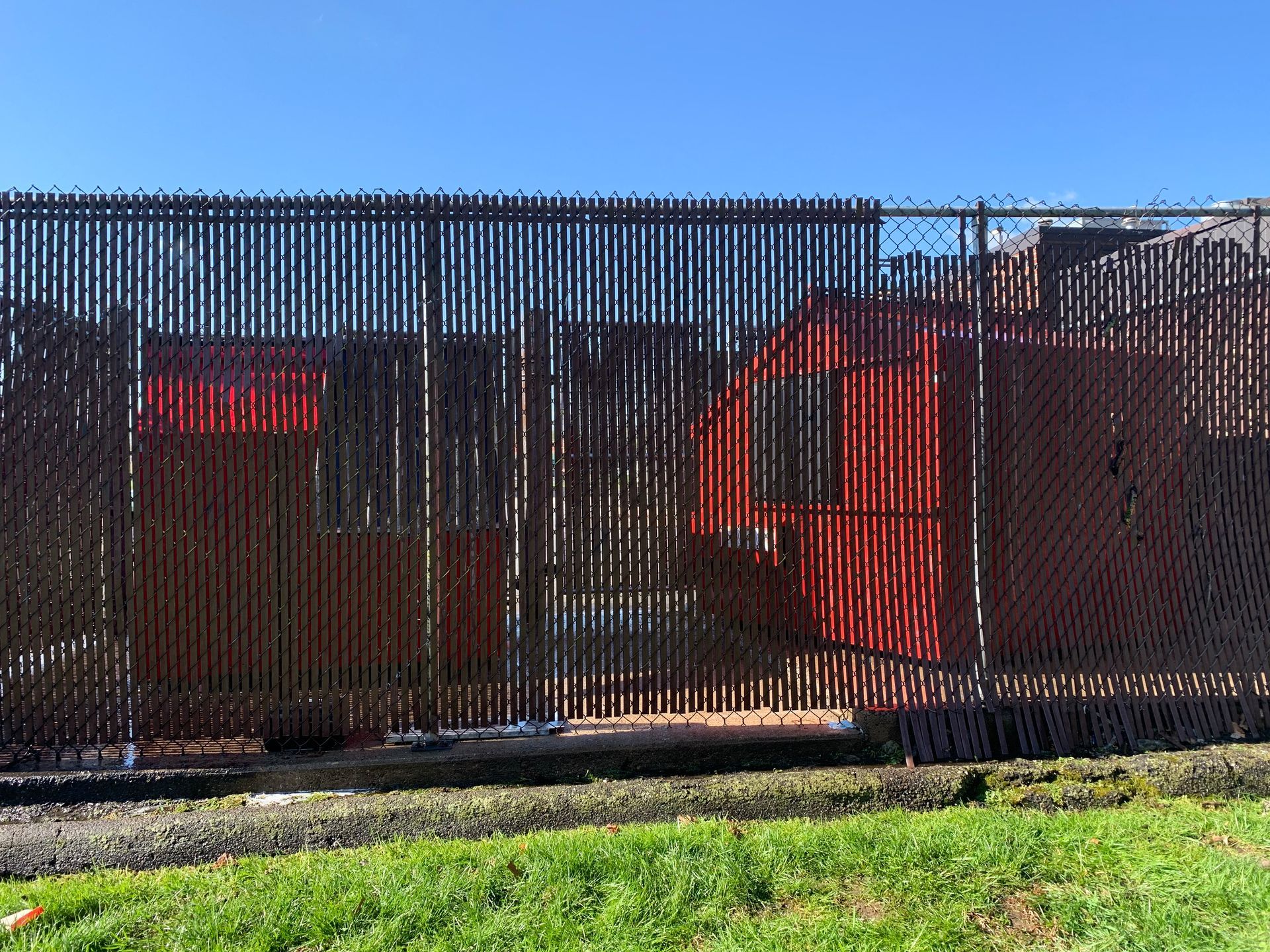 Red structures partially visible behind a dark privacy fence, set against a blue sky. Green grass in the foreground.