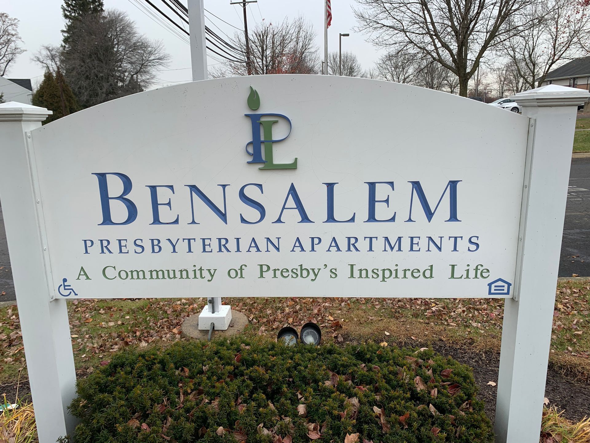 Sign for Bensalem Presbyterian Apartments, white with blue text and logo, green shrubbery in front.
