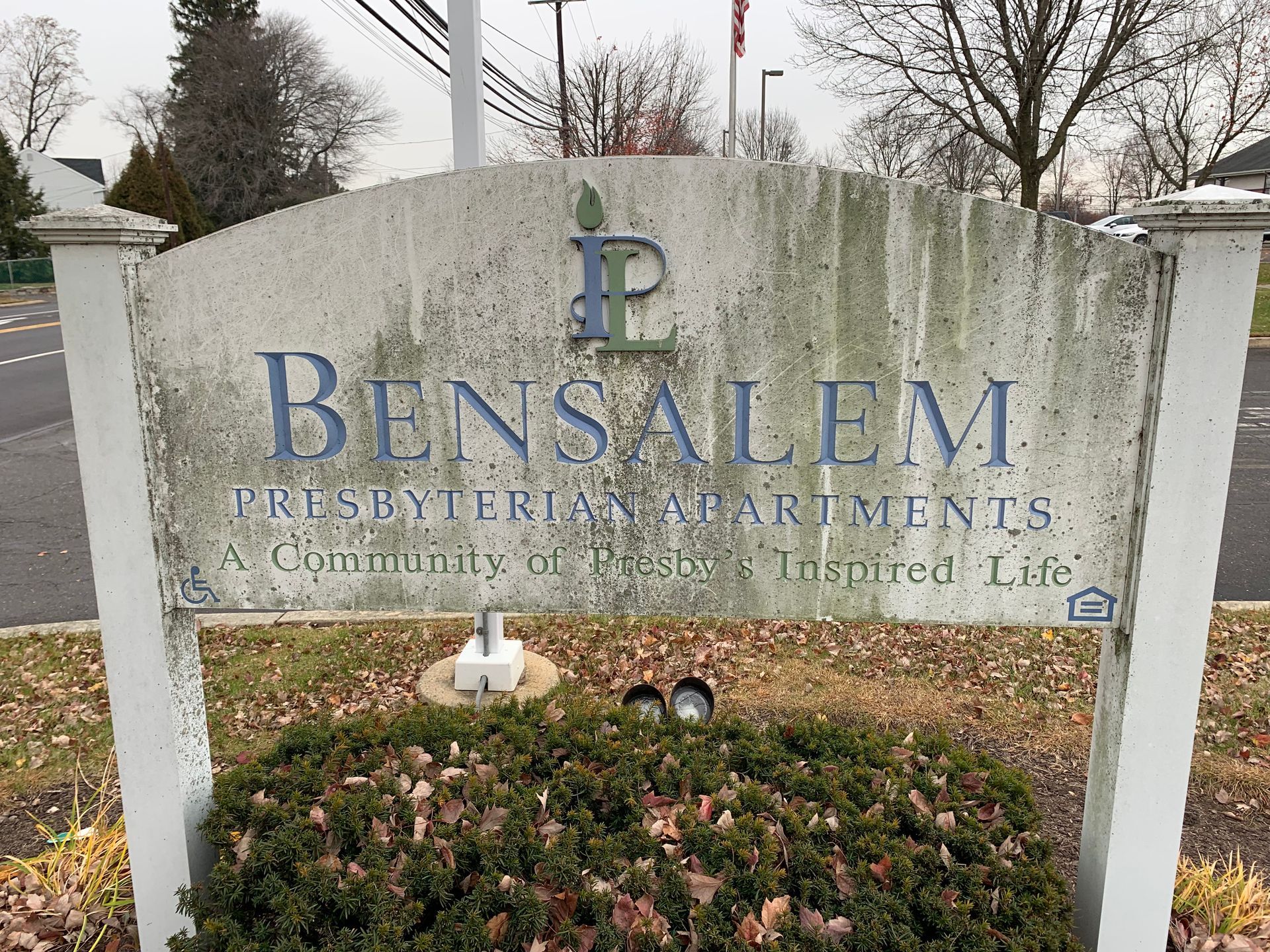 Sign for Bensalem Presbyterian Apartments, weathered with text and logo, atop a small bush.