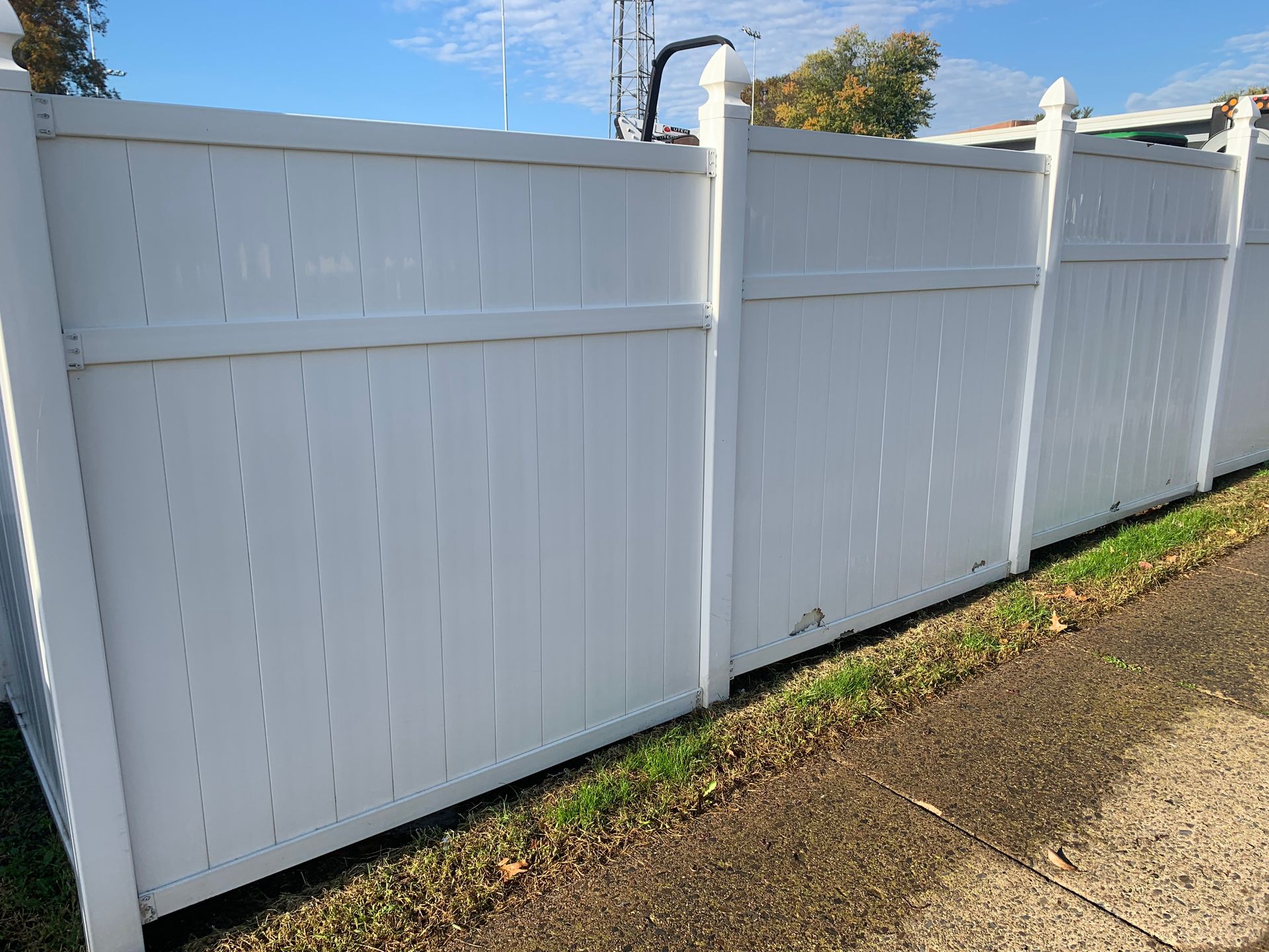 White vinyl fence along a sidewalk with small patches of grass and some leaves.