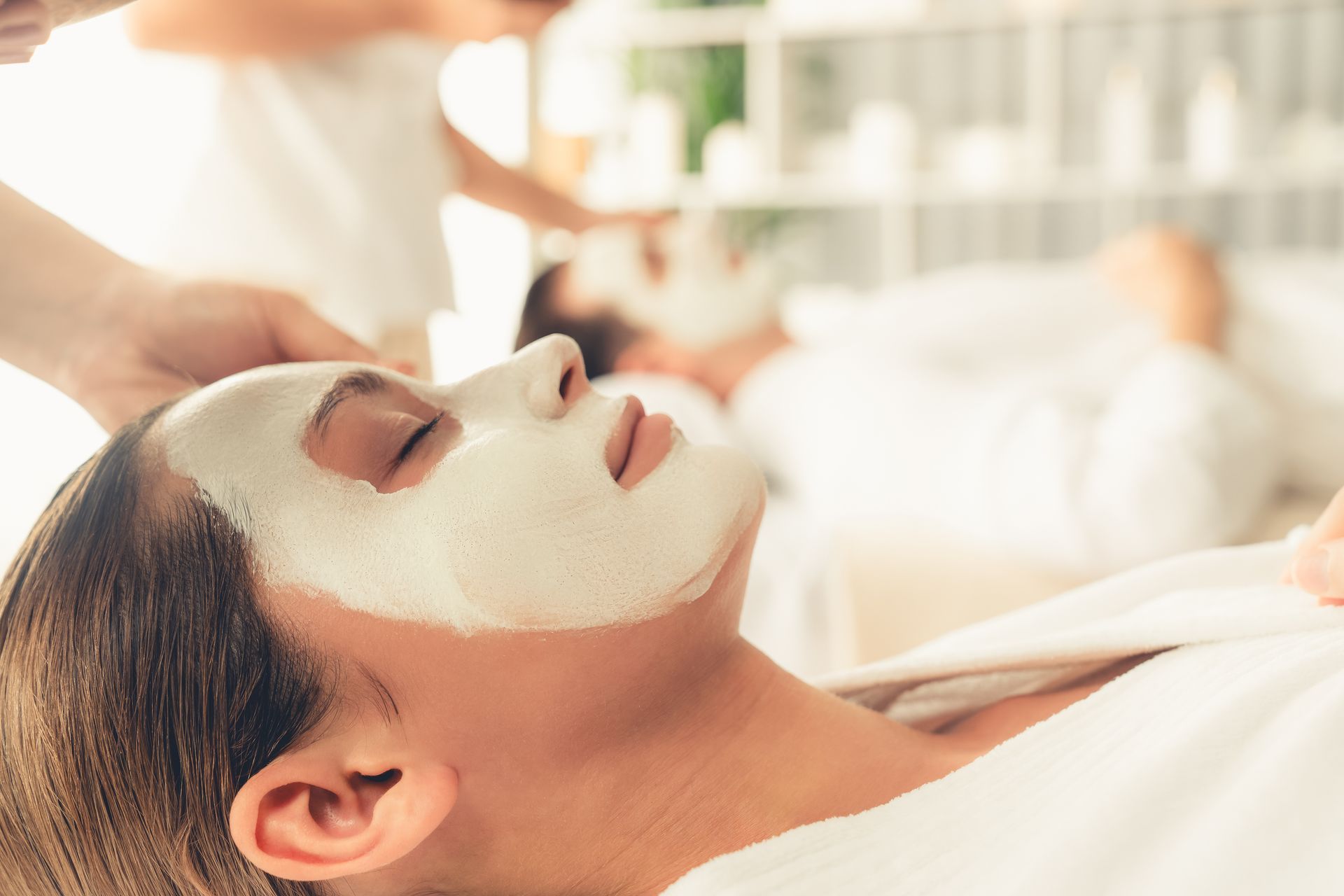 Woman with a white facial mask lying on a spa bed while a therapist applies treatment