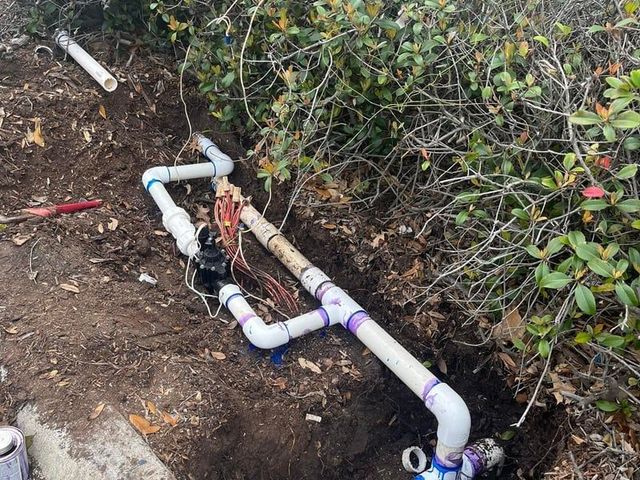 White PVC pipes and valves installed in a dirt trench beside green shrubs.