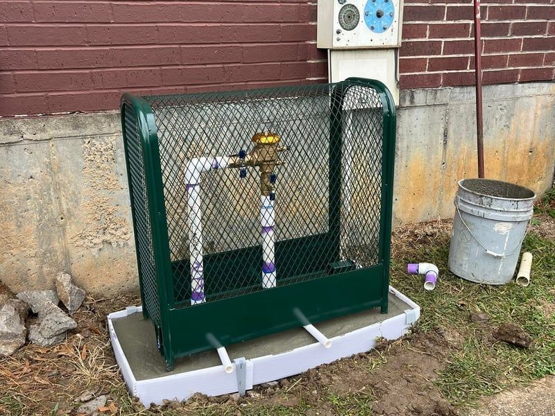 Green metal gas meter enclosure beside brick building with exposed white pipes and a bucket on the ground