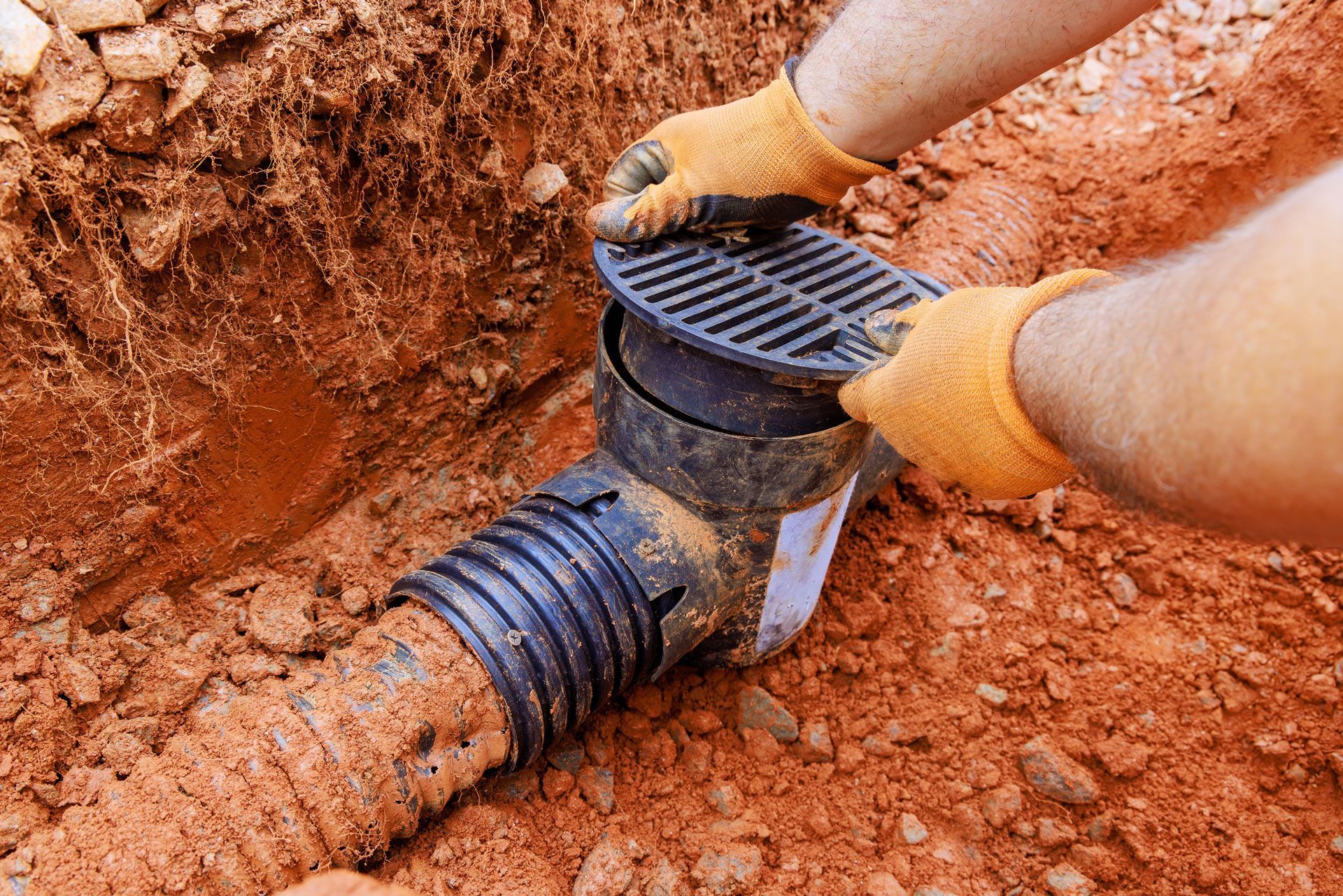 Gloved hands installing a black drainage pipe in red dirt