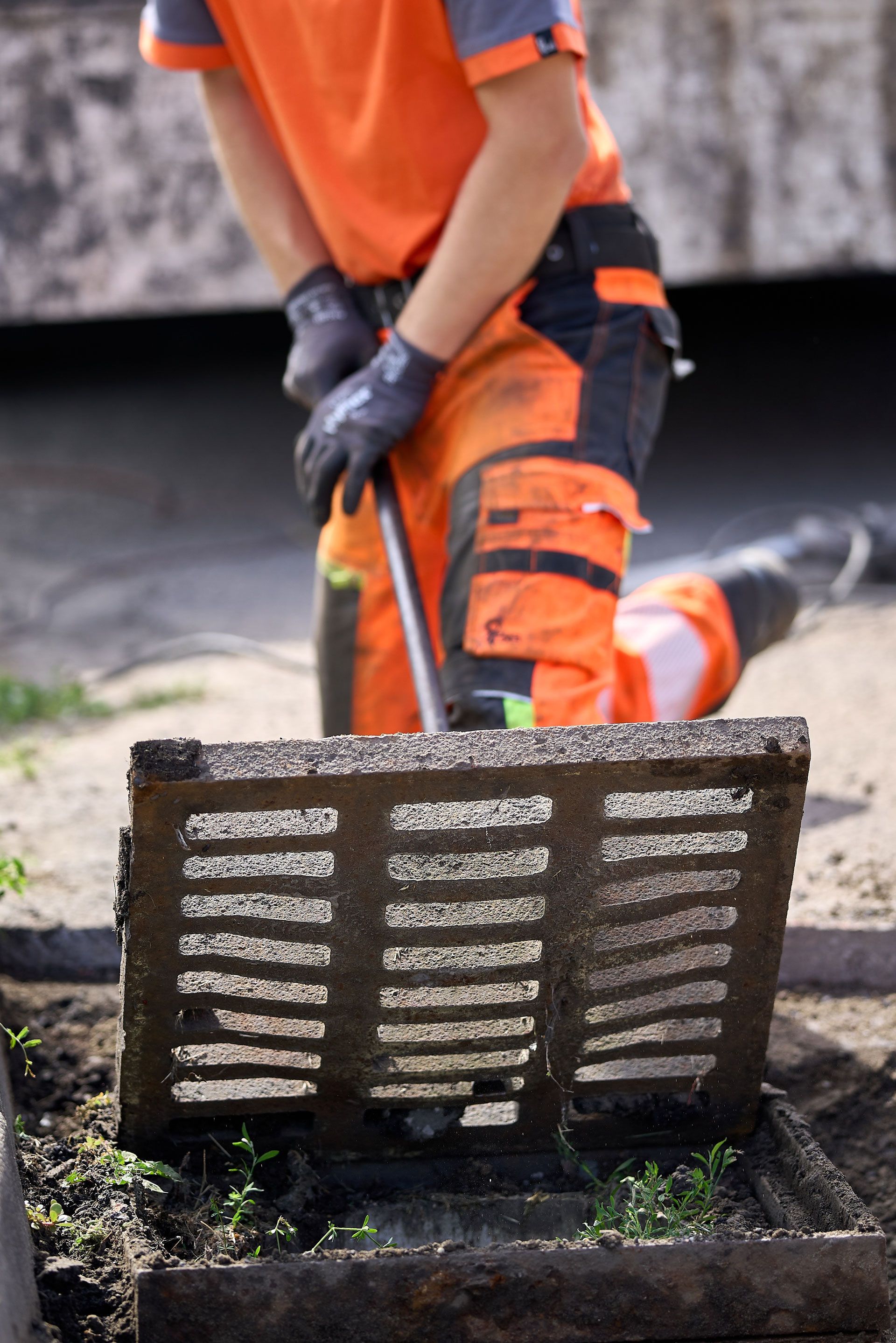 Worker in orange safety gear lifting a metal drain grate at a construction site.