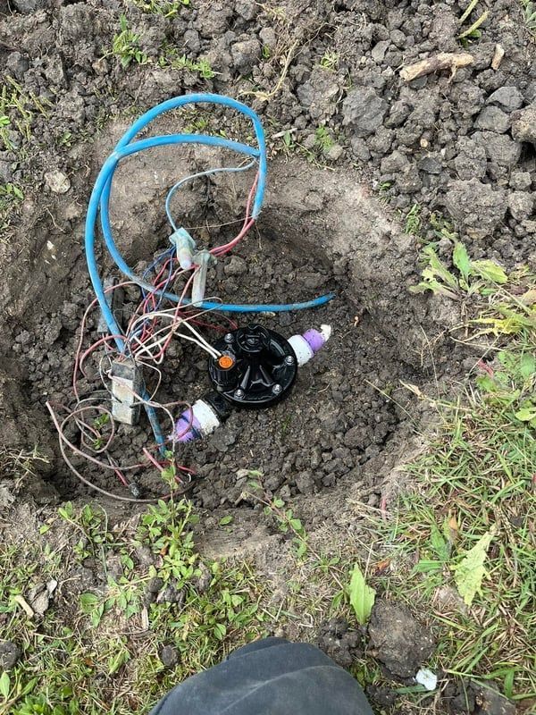 Blue hoses and wiring exposed in a dirt hole outdoors, with a black circular component and purple connectors.