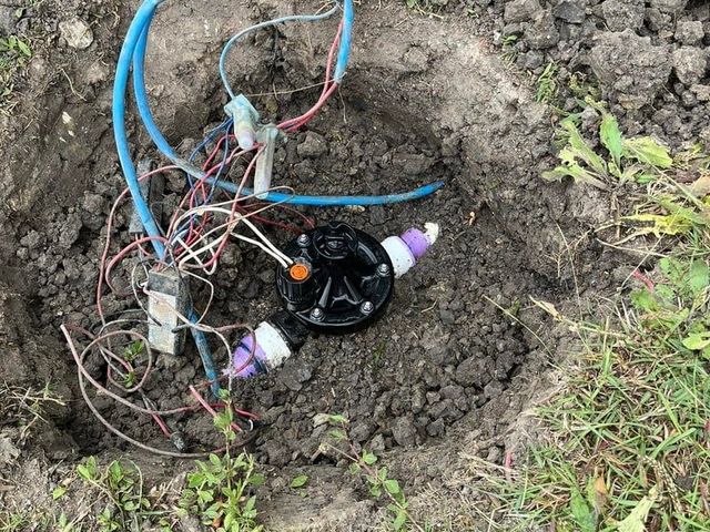 Open muddy hole with exposed wires, blue tubing, and a black plastic device in the ground