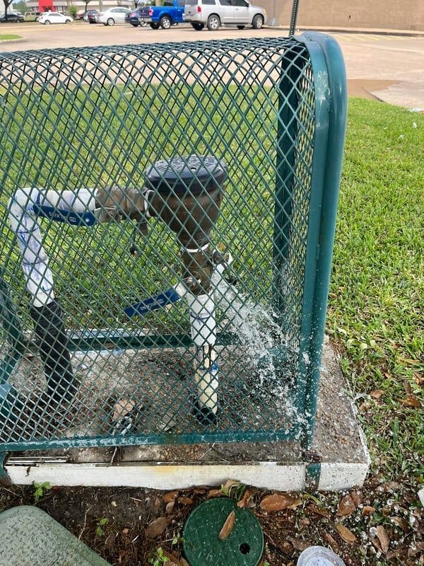 White-and-blue water pipe with black valve inside a green chain-link enclosure beside a sidewalk.