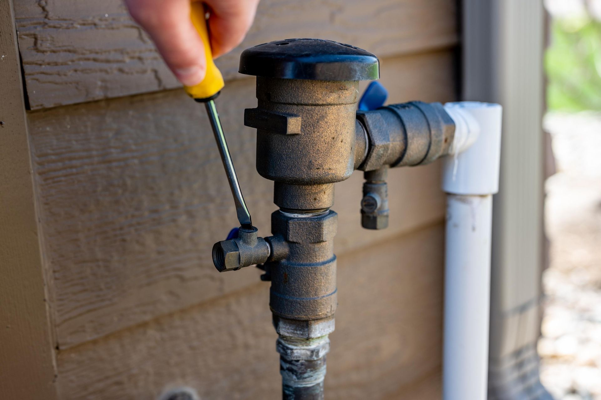 Hand turning a black outdoor water valve beside white PVC pipes on a house exterior