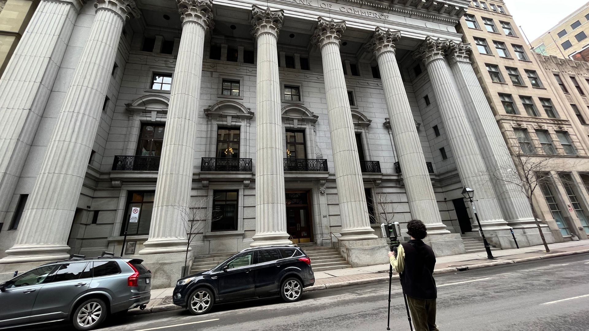 Classical stone building with tall columns, parked cars, and a person standing on the sidewalk below