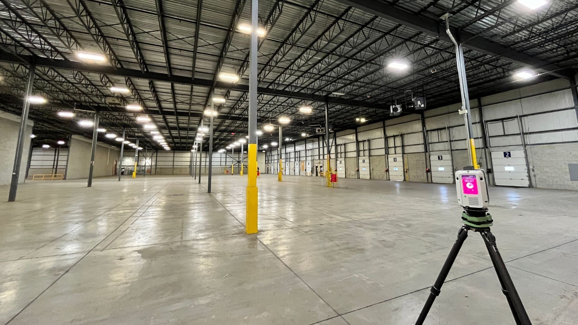 Empty warehouse interior with concrete floor, exposed beams, yellow columns, and a tripod device in the foreground.