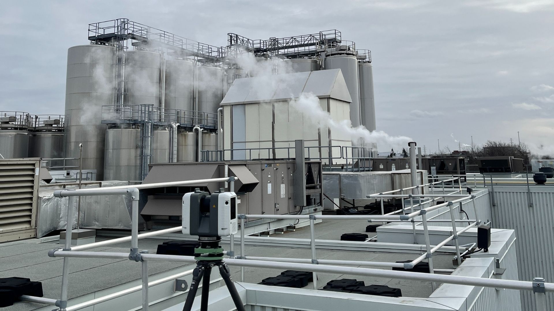 Industrial tanks and equipment on a snowy rooftop under a gray sky, with a tripod in the foreground