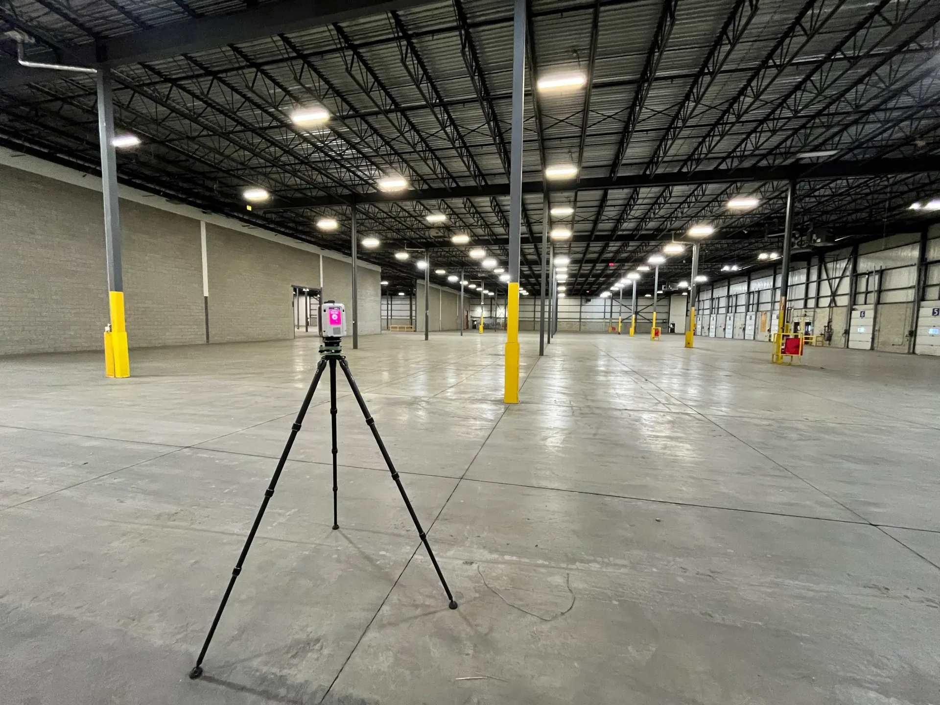 Tripod-mounted device in empty warehouse with high ceiling, gray floor, yellow support poles.