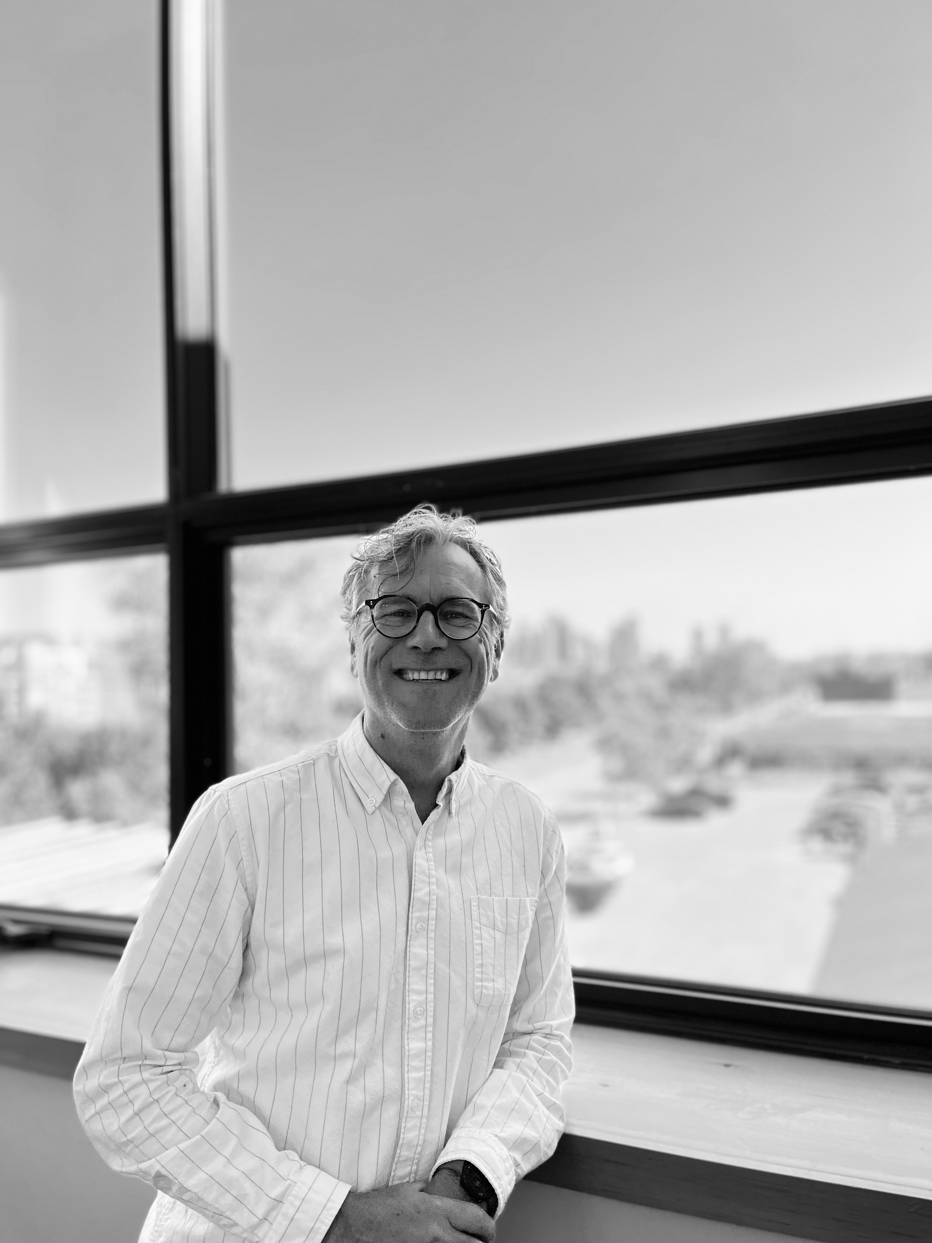 Smiling person in a white shirt standing by a large window with a bright outdoor view