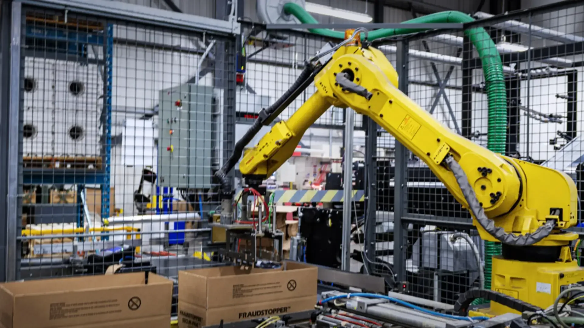 Yellow robotic arm in a factory environment places an item into a cardboard box.