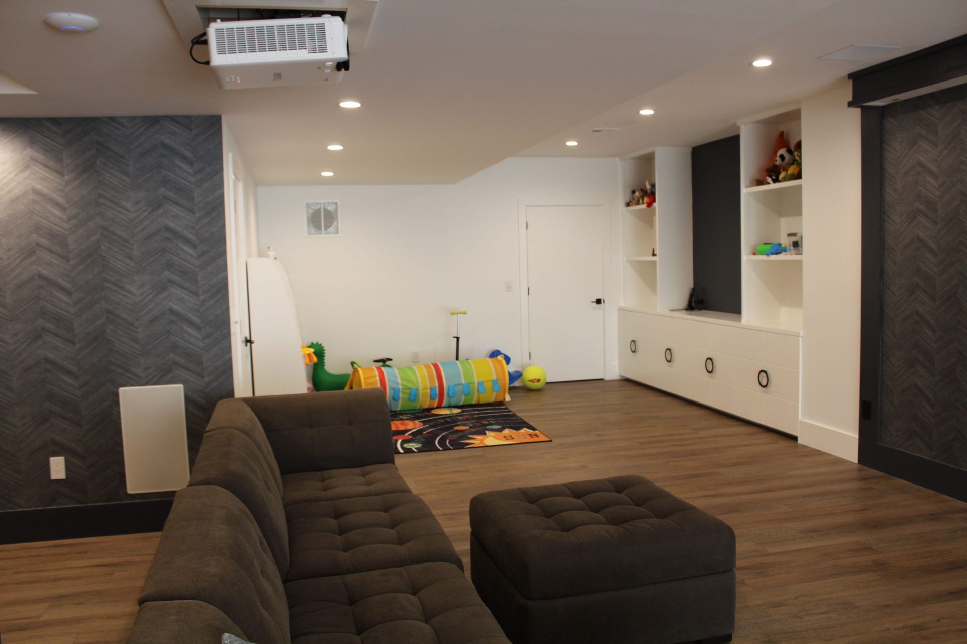 Basement lounge with sectional sofa, built-in shelves, and children’s play tunnel in bright colors.