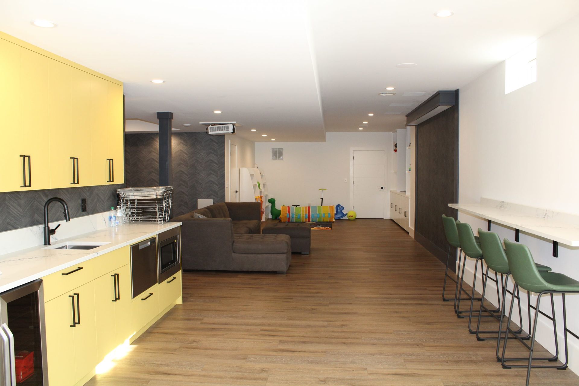 A modern basement kitchen and entertainment area featuring yellow cabinets, a white countertop, and green bar stools.