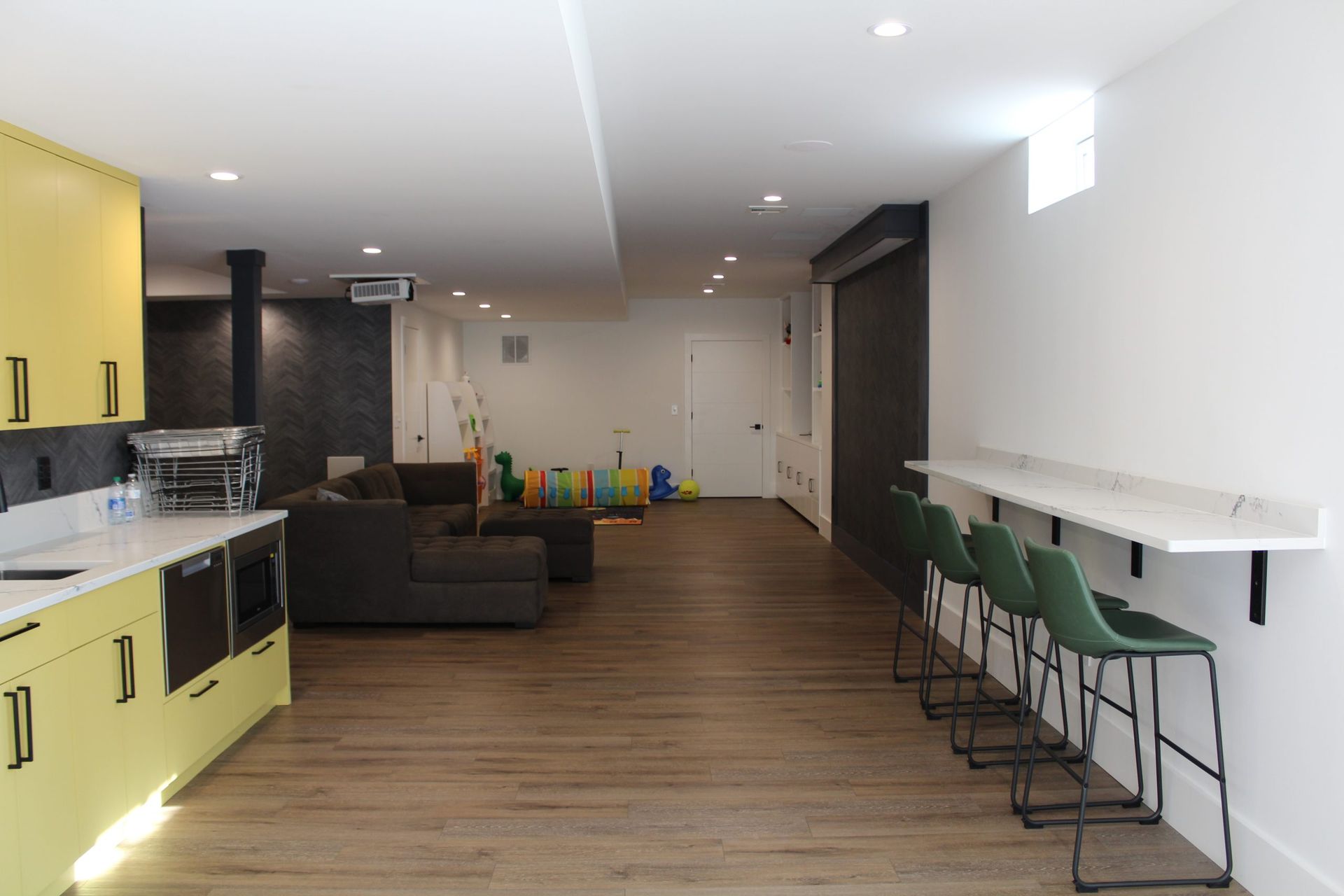 Basement with a kitchen, seating area, and bar. Yellow cabinets contrast with white countertops and wood flooring.