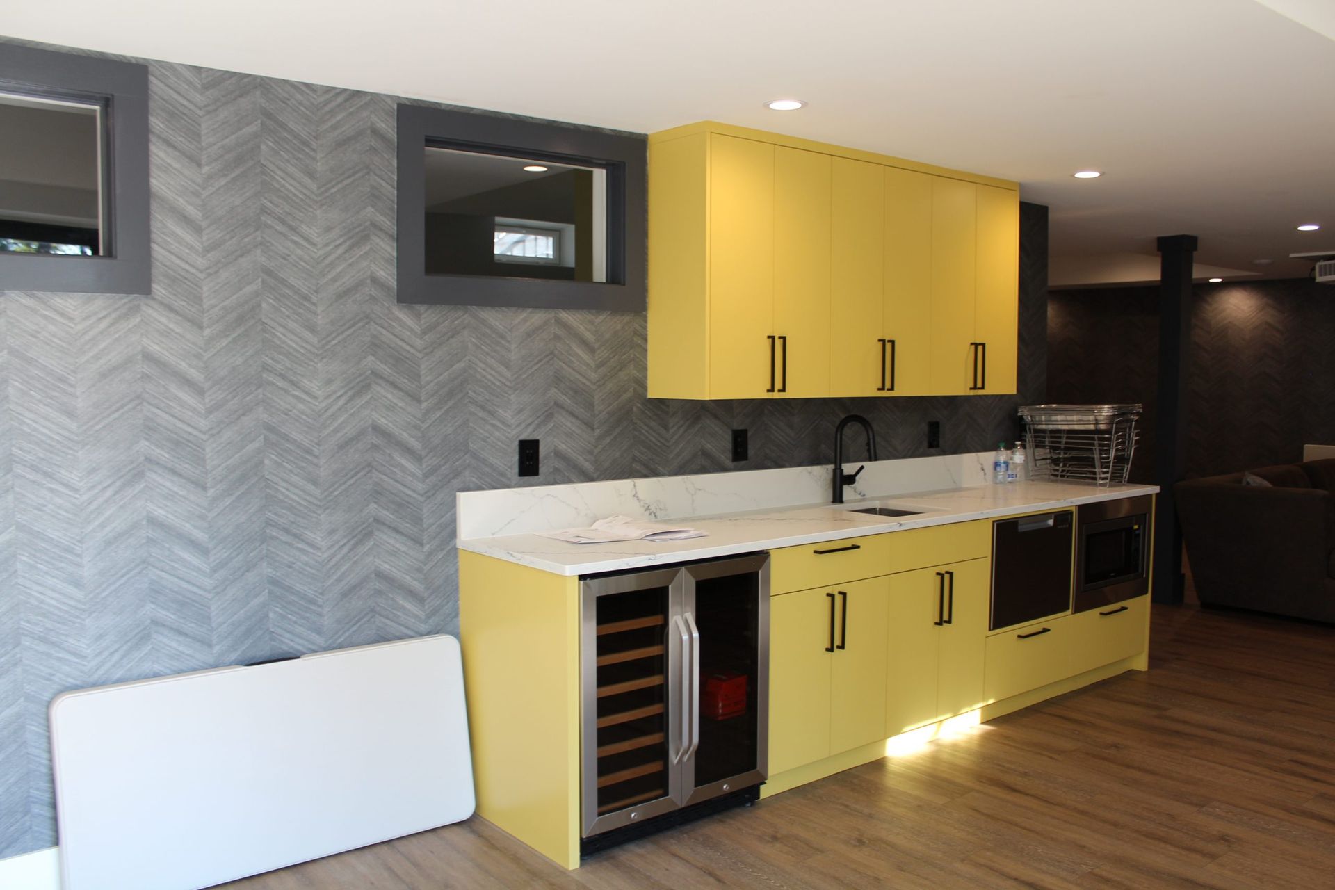 Yellow kitchen cabinets and countertops in a basement with a wine fridge, sink, and microwave.