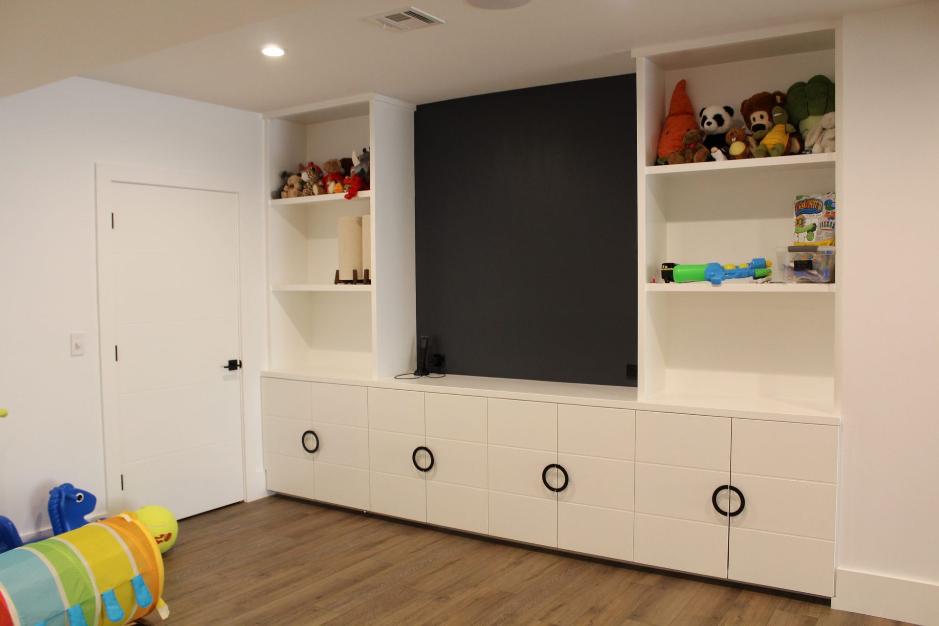 A playroom with built-in white cabinets, shelves with toys, and a large black wall.