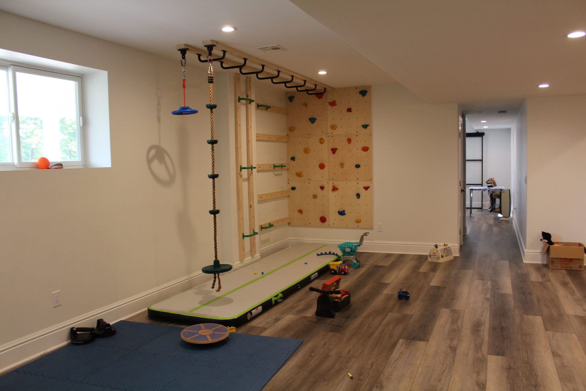 Indoor play area with climbing wall, swing set, and various toys on wooden floors. A child is visible in the adjacent room.