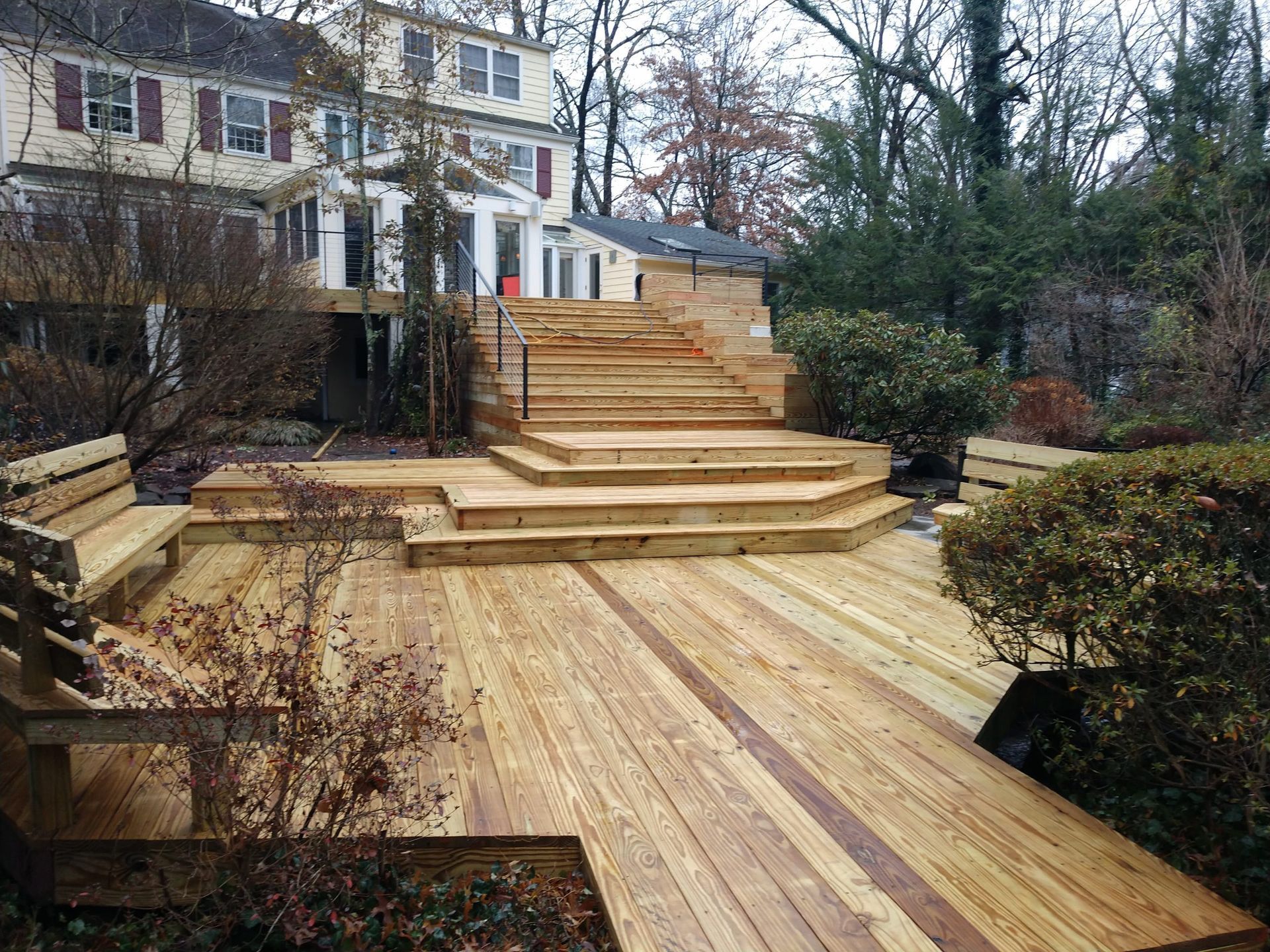 Multi-tiered natural wood deck with built-in benches and stairs surrounded by shrubs.
