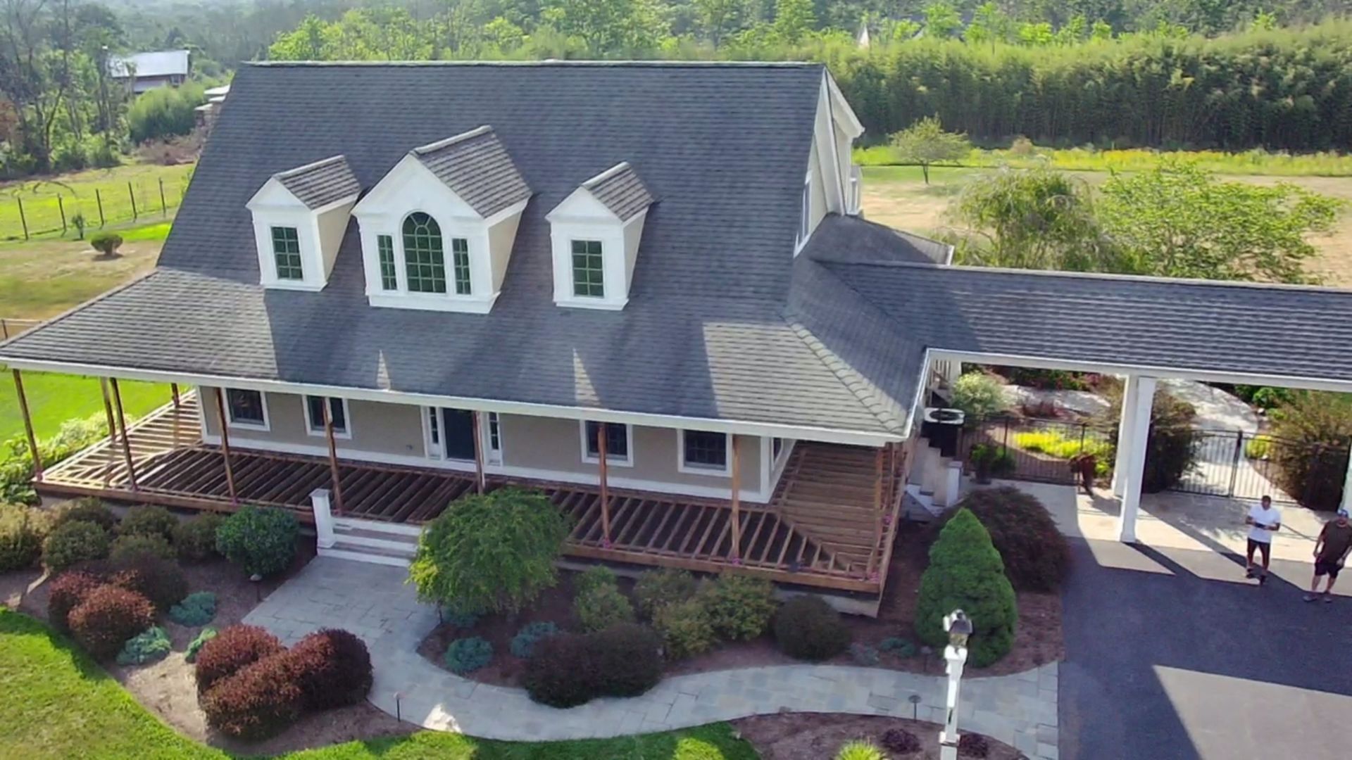 Large house with gray roof and wraparound porch under construction, landscaped yard and driveway.