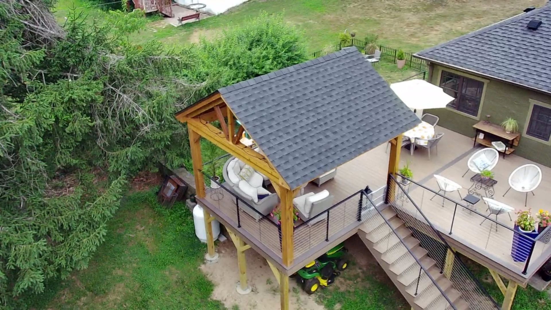 Elevated wooden deck with gable roof, outdoor seating, and stairs, surrounded by lush greenery.