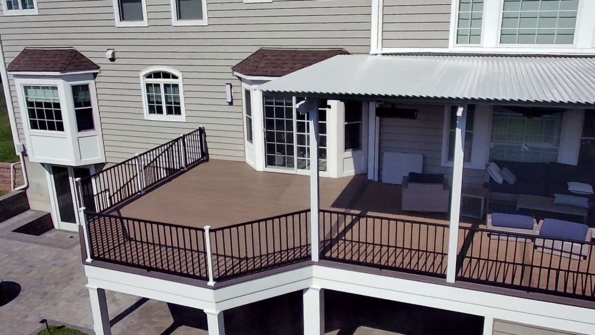 Large back deck with partial roof cover, seating area, and black railing, extending from gray house.