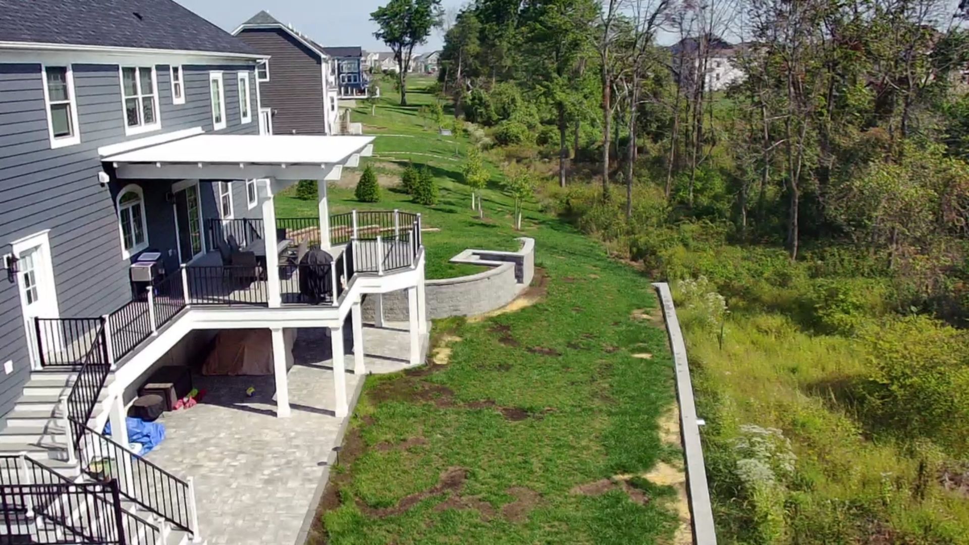 Covered deck with white columns, black railings, and stone patio surrounded by greenery.