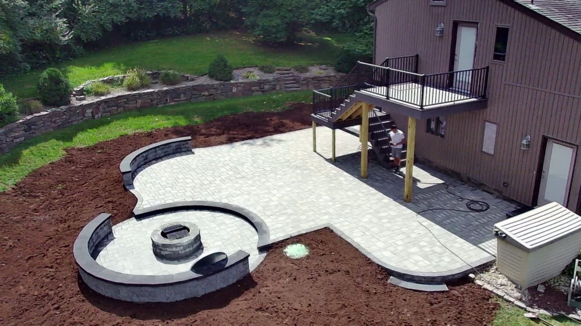 Backyard paver patio with built-in circular fire pit and raised deck with stairs.