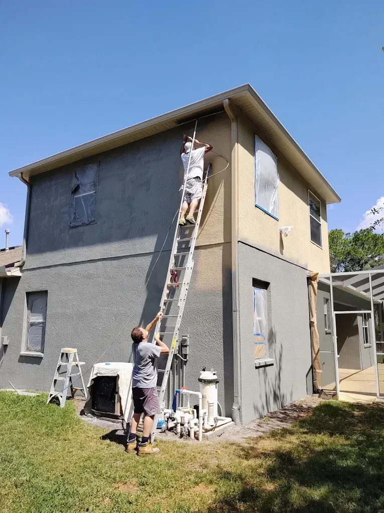 Two men painting a two-story house. One on a ladder, other at its base. Green grass, blue sky.