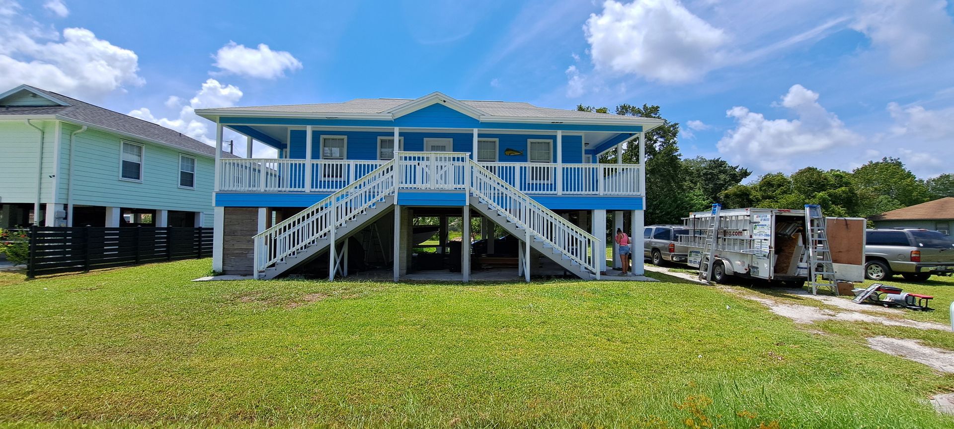Blue house on stilts with wooden stairs, porch, and a grassy lawn.