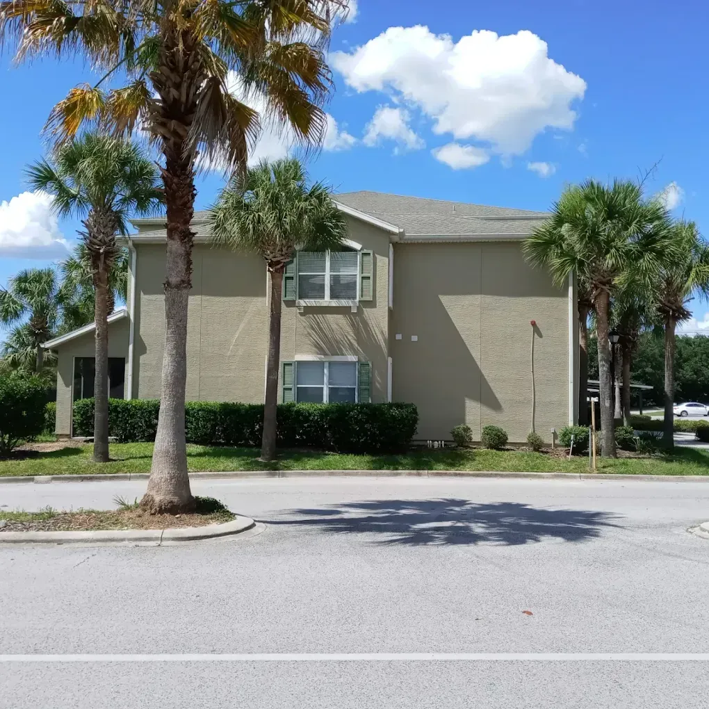 Two-story beige building with palm trees and green hedges, under a blue sky with clouds.