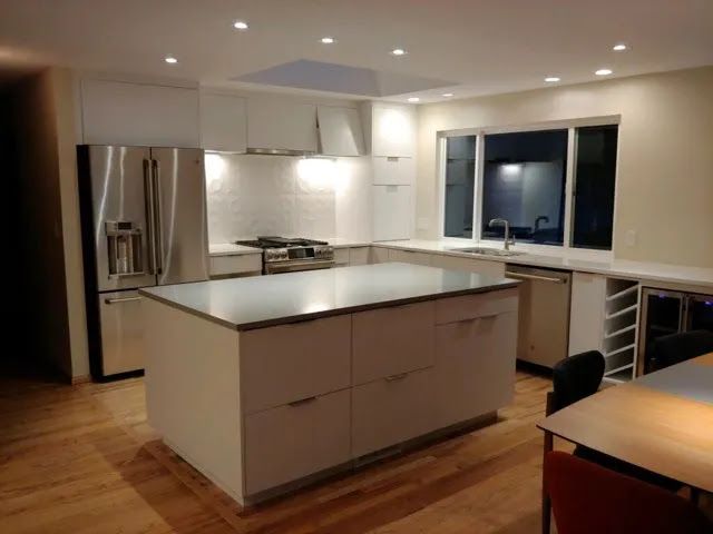 Modern kitchen with white island, stainless fridge, wood floor, and large window at night