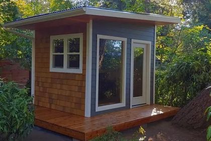 Small modern wooden shed with white trim and large windows, set among trees in a garden